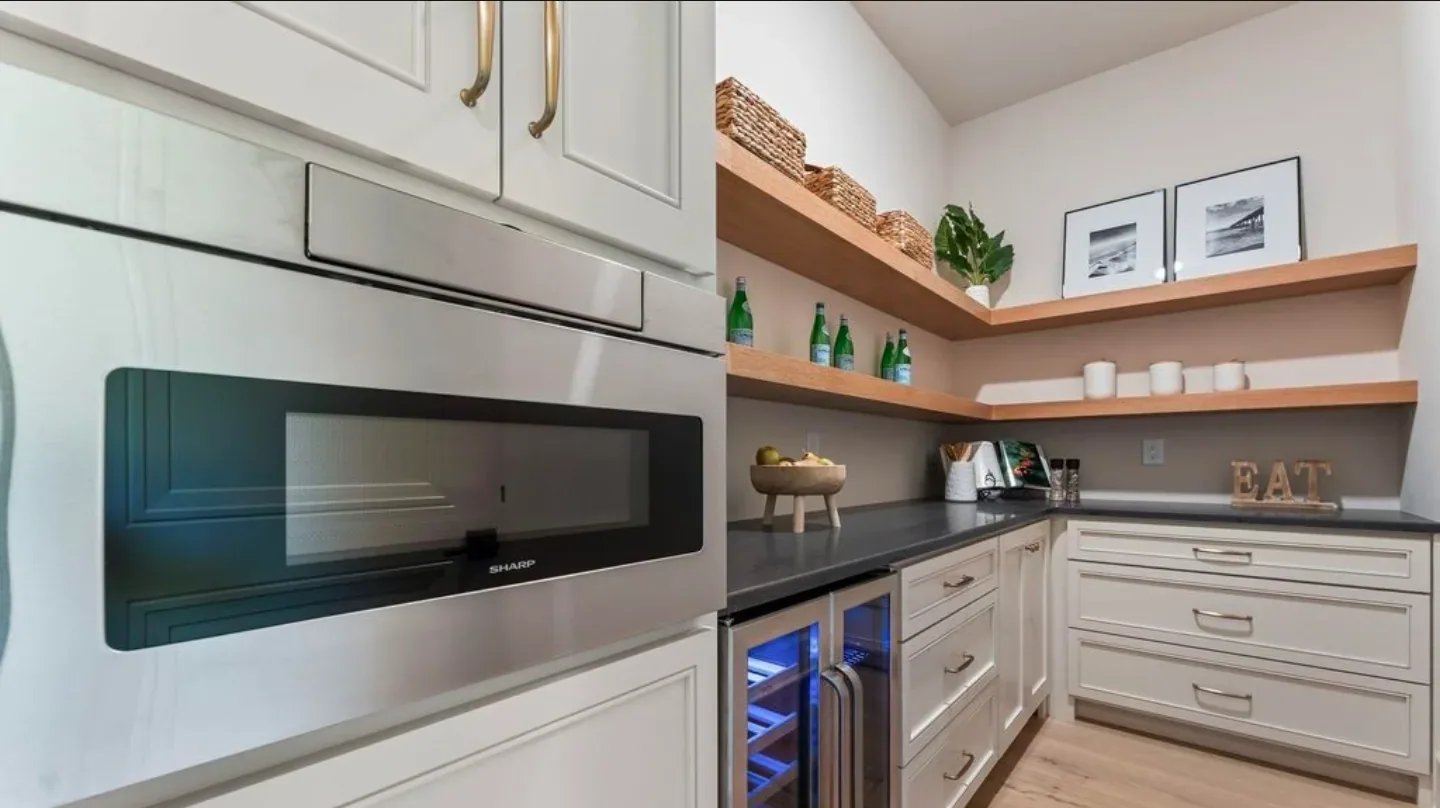 A butler pantry with white shaker drawers, a wine fridge, dark counters and open oak shelves